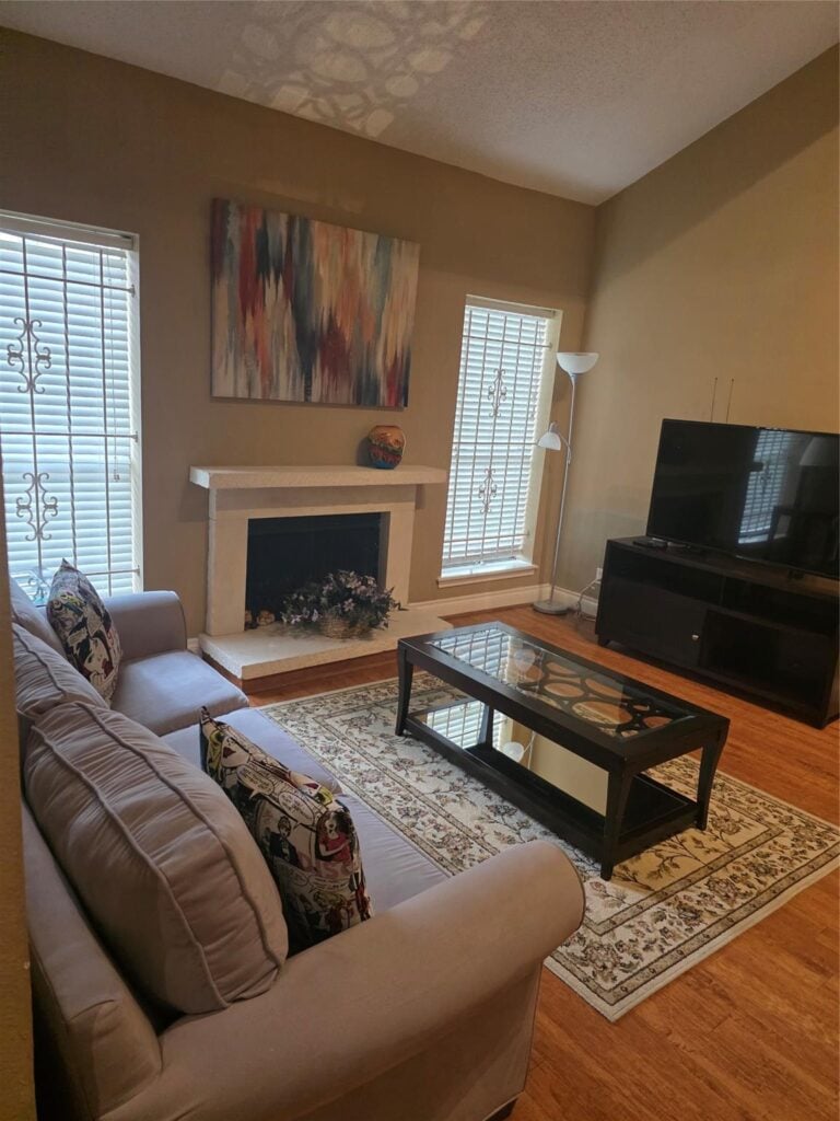 Cozy living room featuring a light gray sofa with decorative pillows, a glass coffee table on an area rug, a fireplace with a decorative piece, and large windows with blinds, showcasing a warm and inviting atmosphere ideal for family gatherings.