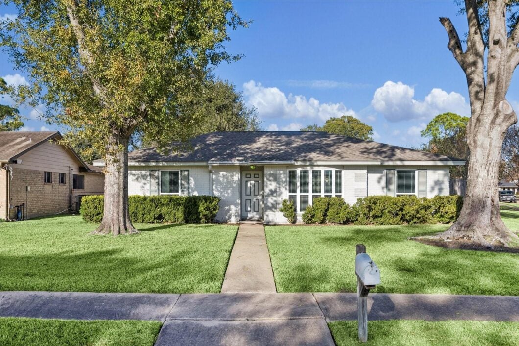 Single-story residential home in Meadows Place, featuring a well-maintained lawn, large trees, and a welcoming entrance, showcasing recent updates and ample parking space.